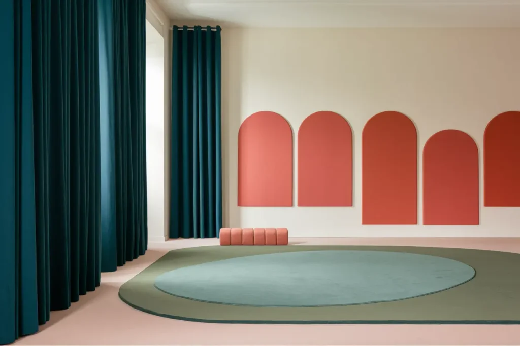 teacher noise sensitivity, A serene classroom scene with deep teal curtains, soft beige walls, and a large sage green rug covering most of the floor. Terra cotta acoustic panels add visual interest on one side, while coral-colored fabric-wrapped bulletin boards soften the walls.